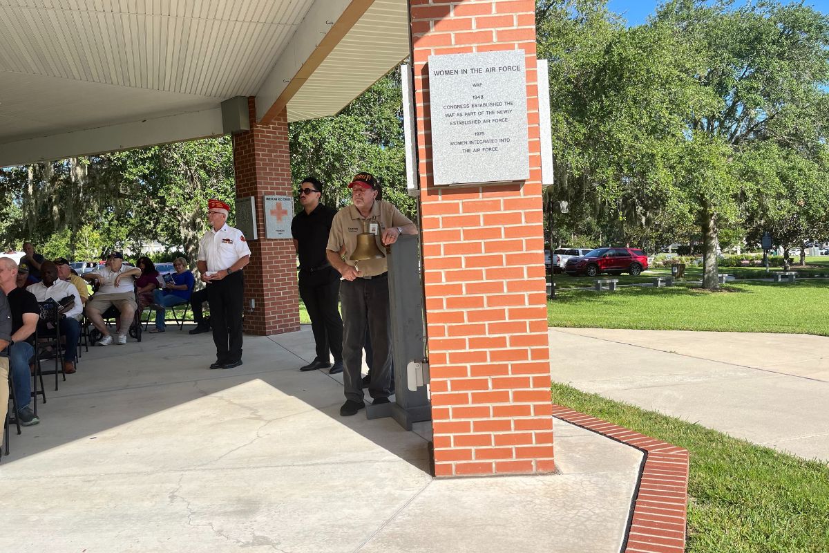 A man dressed in a uniform, stands by a bell, with people to his left and brick column with a granite plaque affixed to it to his right.