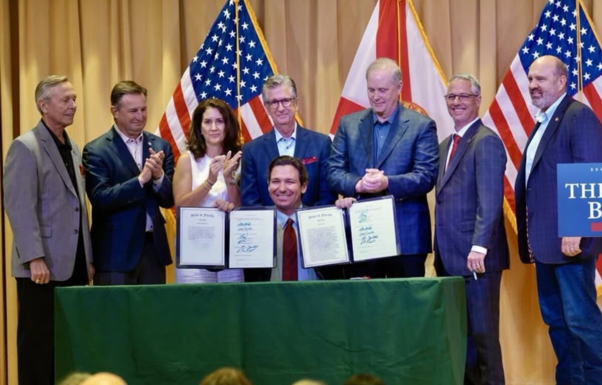Gov. Ron DeSantis smiles while holding signed budget documents surrounded by officials at a ceremony in Wildwood, Florida.