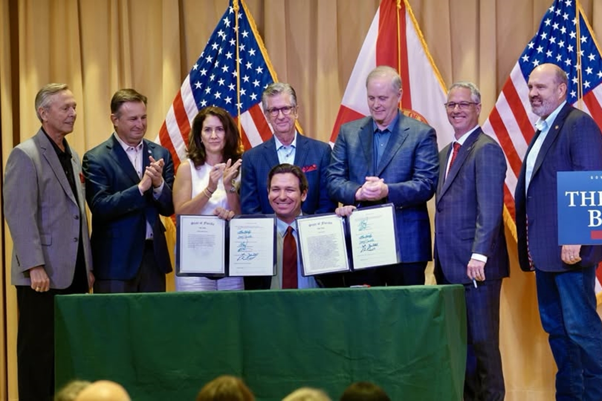 Gov. Ron DeSantis smiles while holding signed budget documents surrounded by officials at a ceremony in Wildwood, Florida.