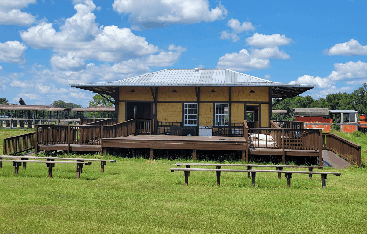 A wooden depot-style building with yellow paneling and a metal roof stands on a grassy field under a bright blue sky with scattered clouds. The building features ramps and a deck with railings, while train cars can be seen in the background.