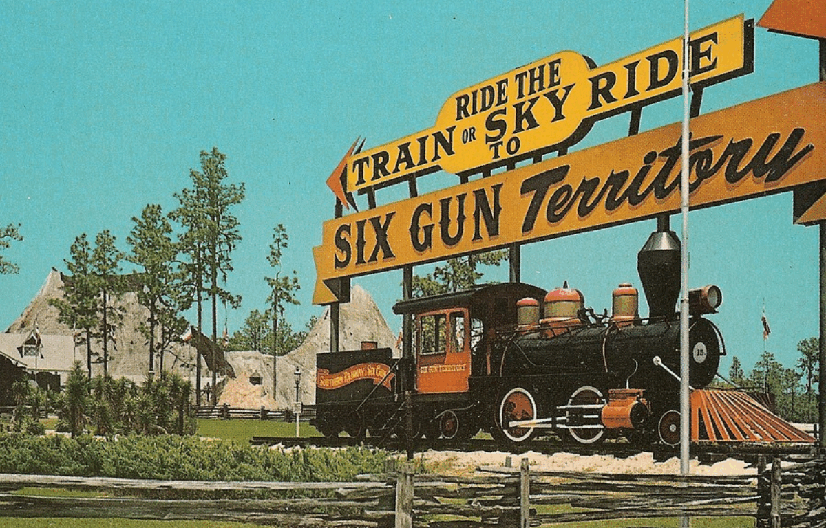 The entrance to Six Gun Territory. A steam locomotive sits on a piece of train track with a sign that reads "Ride the Train or Sky Ride to Six Gun Territory." Trees and a mountain landscape sit behind the train in the background while a small portion of fence sits in the foreground.