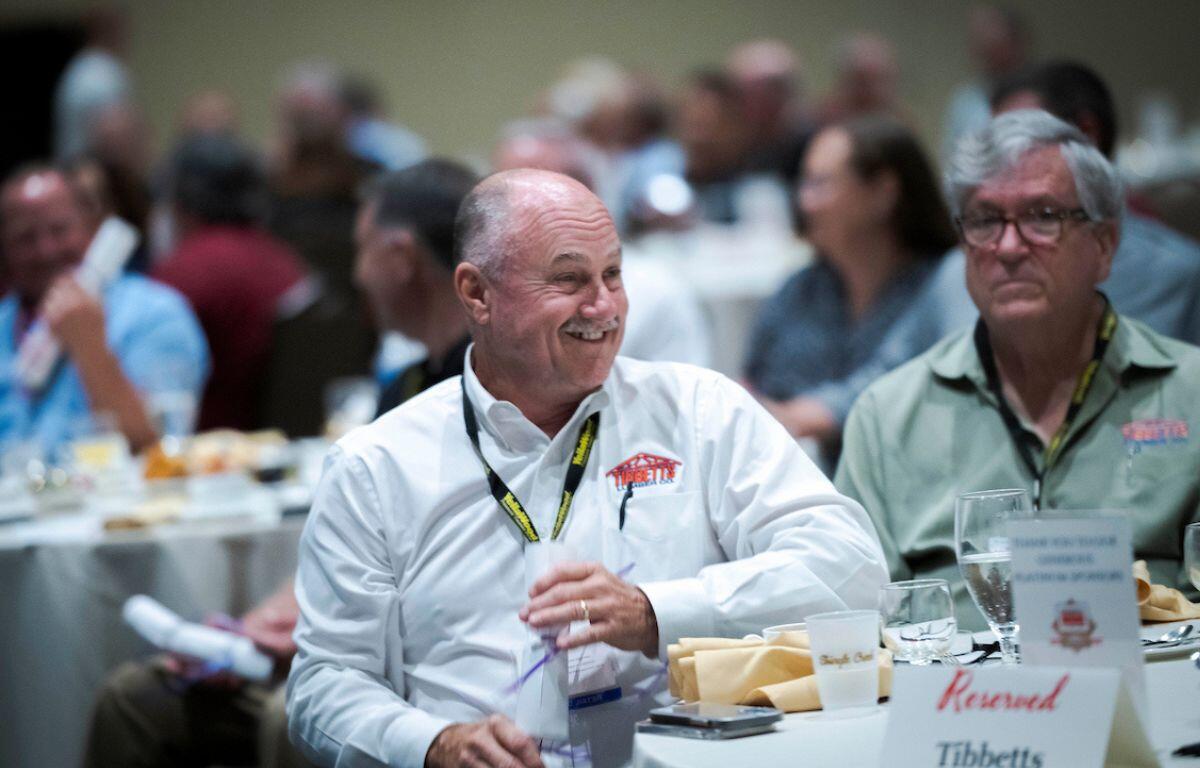 Man smiling and seated at a table during an FBMA event with other attendees in the background.