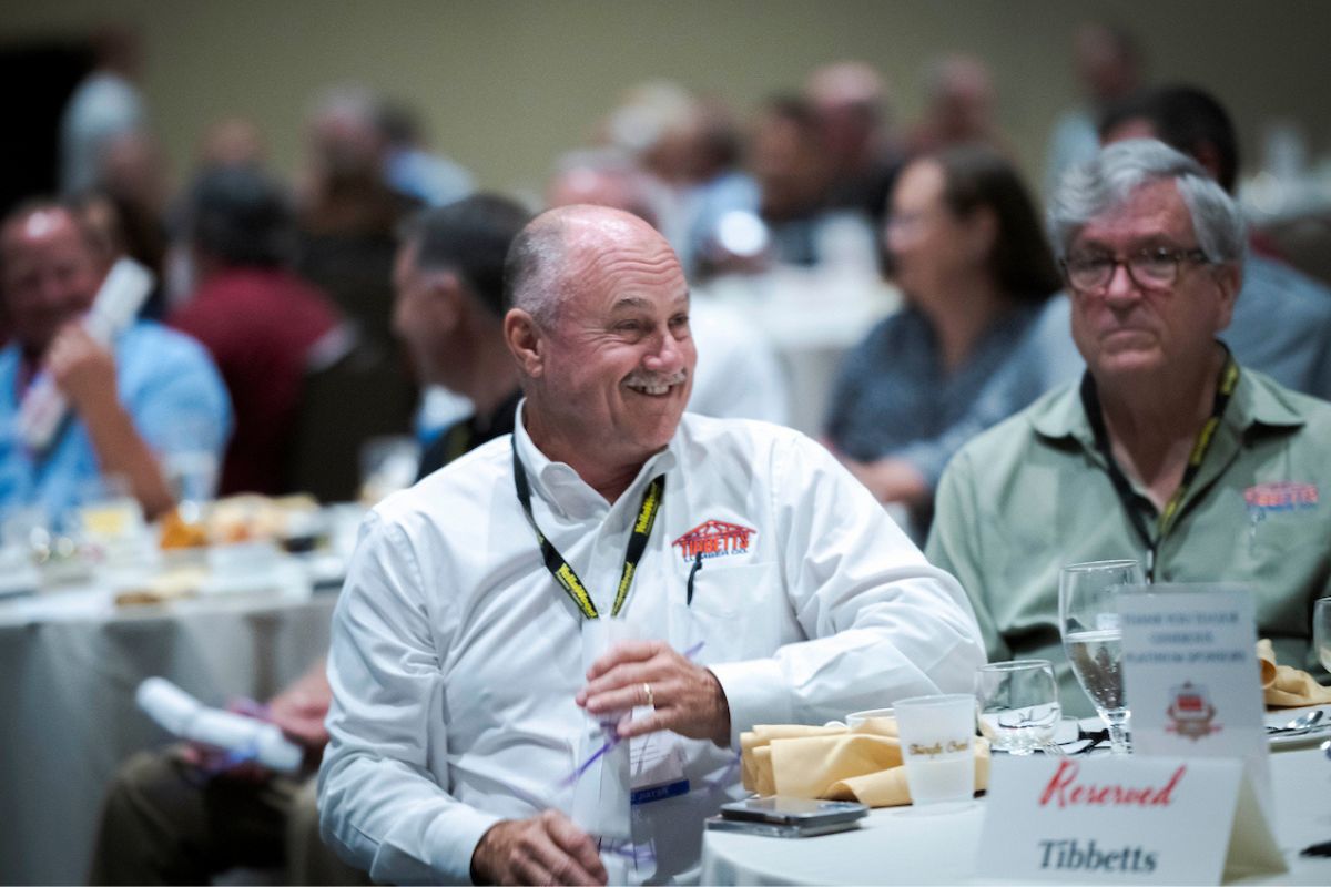 Man smiling and seated at a table during an FBMA event with other attendees in the background.
