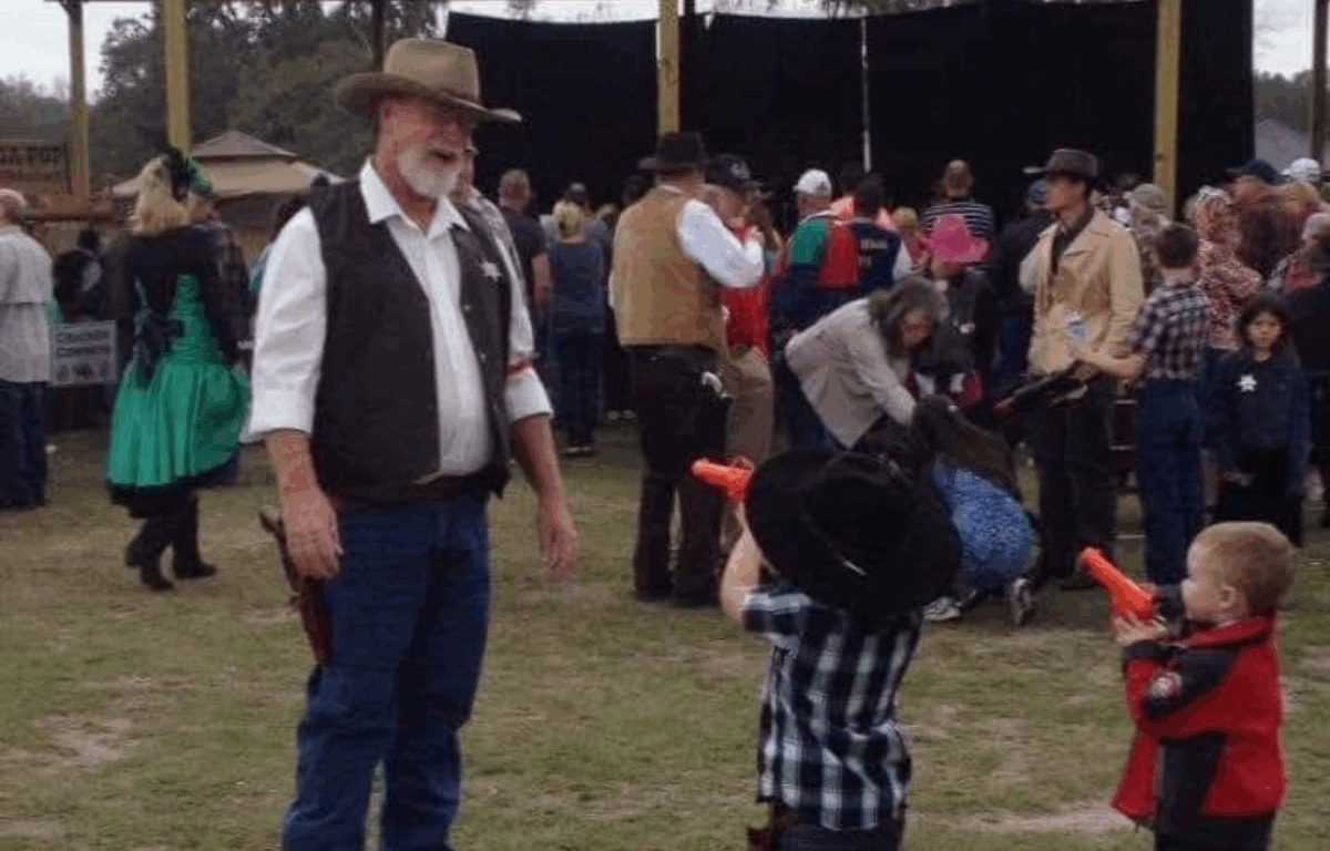 A Western-themed festival scene features a man dressed as a sheriff smiling as two young children, both in cowboy hats, playfully aim toy guns at him. Behind them, adults in frontier attire mingle among a large crowd of attendees.
