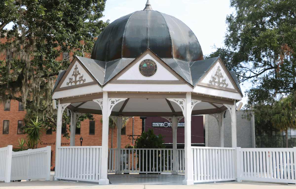 Closeup of Ocala Downtown Gazebo with white support beams and fencing surrounding it and leading down to the square. White triangles surround the top of the gazebo with a blue-gray dome sitting atop it and a City of Ocala circular logo placed center in the front facing triangle.