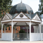 Closeup of Ocala Downtown Gazebo with white support beams and fencing surrounding it and leading down to the square. White triangles surround the top of the gazebo with a blue-gray dome sitting atop it and a City of Ocala circular logo placed center in the front facing triangle.