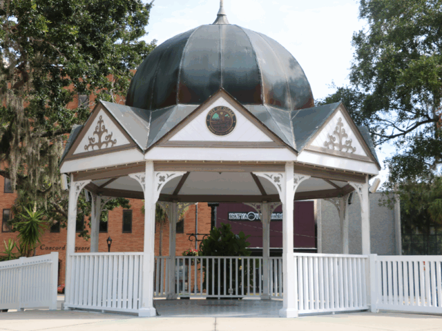 Closeup of Ocala Downtown Gazebo with white support beams and fencing surrounding it and leading down to the square. White triangles surround the top of the gazebo with a blue-gray dome sitting atop it and a City of Ocala circular logo placed center in the front facing triangle.