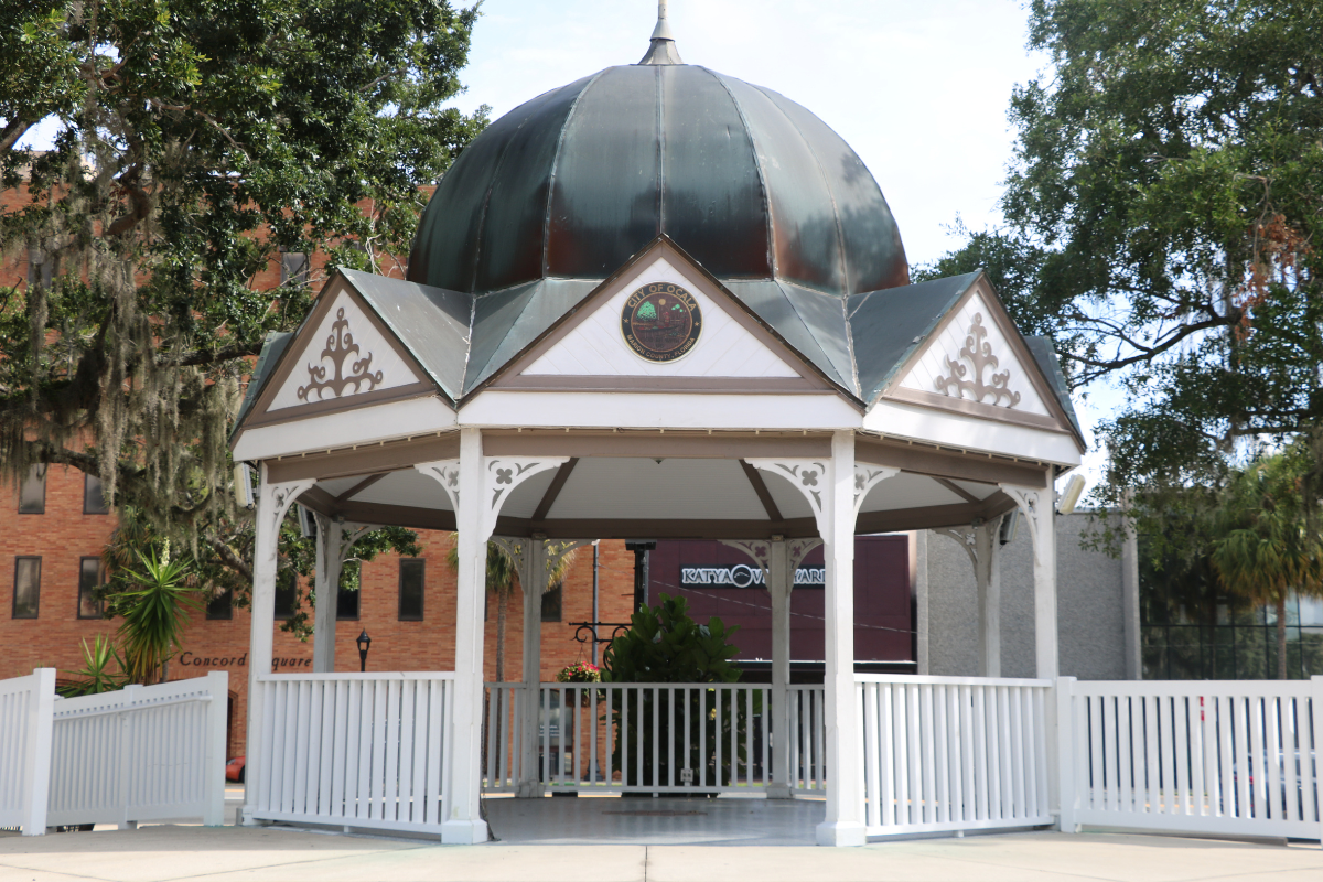 Closeup of Ocala Downtown Gazebo with white support beams and fencing surrounding it and leading down to the square. White triangles surround the top of the gazebo with a blue-gray dome sitting atop it and a City of Ocala circular logo placed center in the front facing triangle.