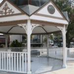 A white gazebo with decorative trim and a metal roof stands near a street corner in Downtown Ocala. The top of the structure features a seal that reads “City of Ocala, Marion County, Florida.” The gazebo is surrounded by a white picket fence and sits on a concrete platform. In the background, there are commercial buildings, parked cars, trees, and street signs.