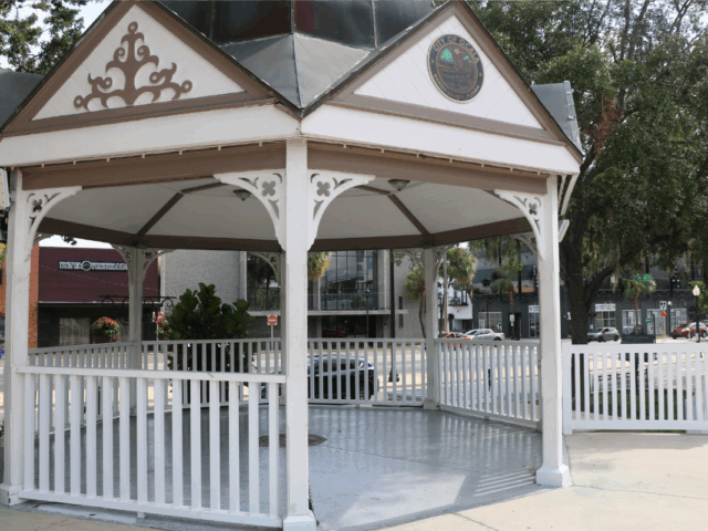 A white gazebo with decorative trim and a metal roof stands near a street corner in Downtown Ocala. The top of the structure features a seal that reads “City of Ocala, Marion County, Florida.” The gazebo is surrounded by a white picket fence and sits on a concrete platform. In the background, there are commercial buildings, parked cars, trees, and street signs.