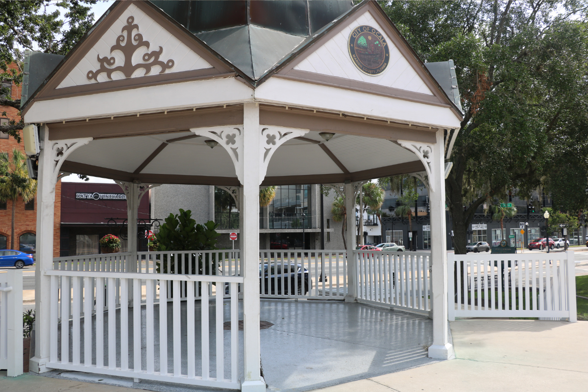 A white gazebo with decorative trim and a metal roof stands near a street corner in Downtown Ocala. The top of the structure features a seal that reads “City of Ocala, Marion County, Florida.” The gazebo is surrounded by a white picket fence and sits on a concrete platform. In the background, there are commercial buildings, parked cars, trees, and street signs.