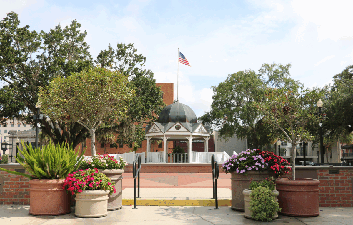 The Ocala Downtown Square front entrance featuring a white gazebo with a blue-gray dome roof and an American flag standing above it, lifted on a platform and surrounded by trees on either side. The foreground includes various planters with a colorful array of flowers and small trees with short brick walls on either side and black rails leading up to the town square and gazebo.