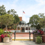 The Ocala Downtown Square front entrance featuring a white gazebo with a blue-gray dome roof and an American flag standing above it, lifted on a platform and surrounded by trees on either side. The foreground includes various planters with a colorful array of flowers and small trees with short brick walls on either side and black rails leading up to the town square and gazebo.