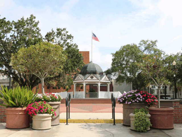 The Ocala Downtown Square front entrance featuring a white gazebo with a blue-gray dome roof and an American flag standing above it, lifted on a platform and surrounded by trees on either side. The foreground includes various planters with a colorful array of flowers and small trees with short brick walls on either side and black rails leading up to the town square and gazebo.