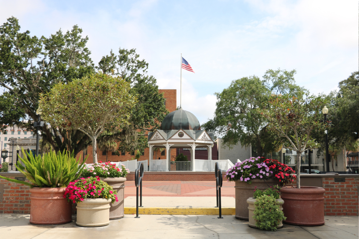 The Ocala Downtown Square front entrance featuring a white gazebo with a blue-gray dome roof and an American flag standing above it, lifted on a platform and surrounded by trees on either side. The foreground includes various planters with a colorful array of flowers and small trees with short brick walls on either side and black rails leading up to the town square and gazebo.