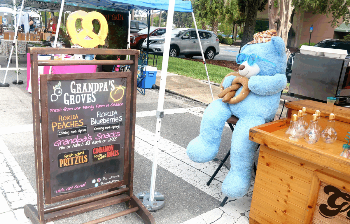 A food vendor advertises food on a chalkboard sign. The vendor, Grandpa's Grove, offers baked pretzels and cinnamon rolls with a blue teddy bear with a pretzel sit on a stood greeting customers.