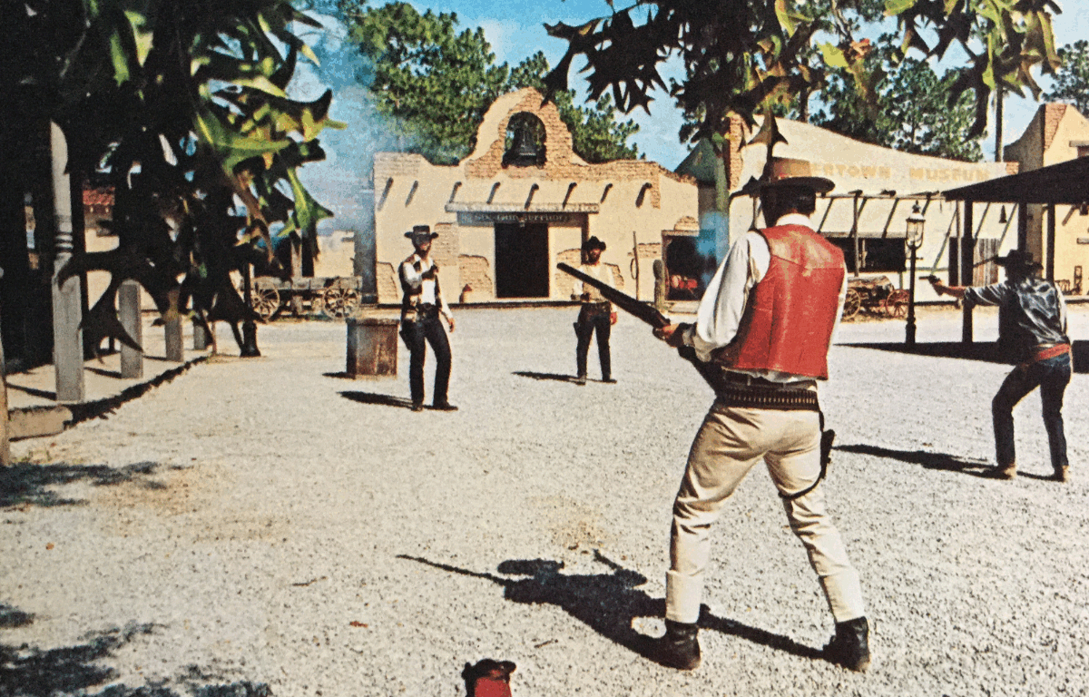 A mock Western shootout occurring in a open-air field of gravel and is surrounded by themed architecture. Four cowboys are holding various models of firearms with branches in the foreground.