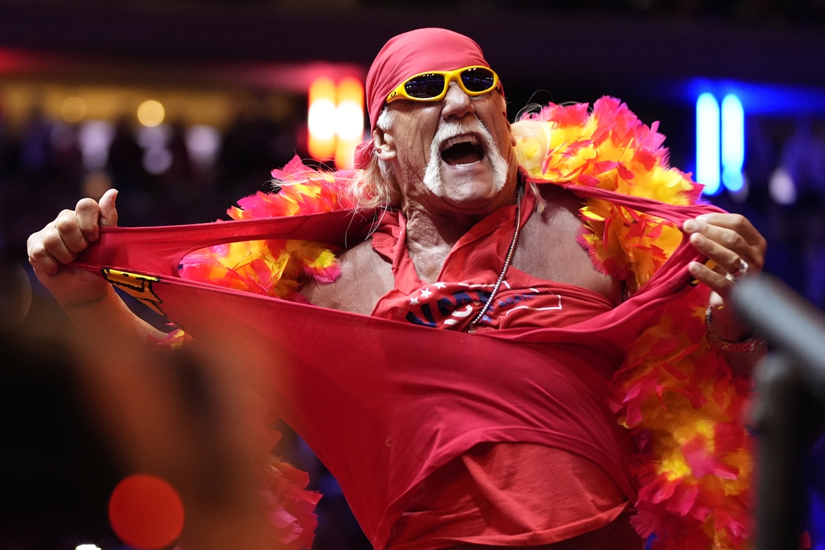 Hulk Hogan tears his shirt while performing in a wrestling arena, wearing a red bandana, yellow sunglasses, and a feathered boa.