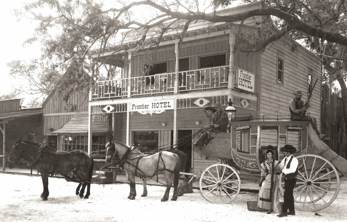 A horse-drawn carriage sits in front of the Frontier Hotel, a two-story building sitting on a street corner in the park.