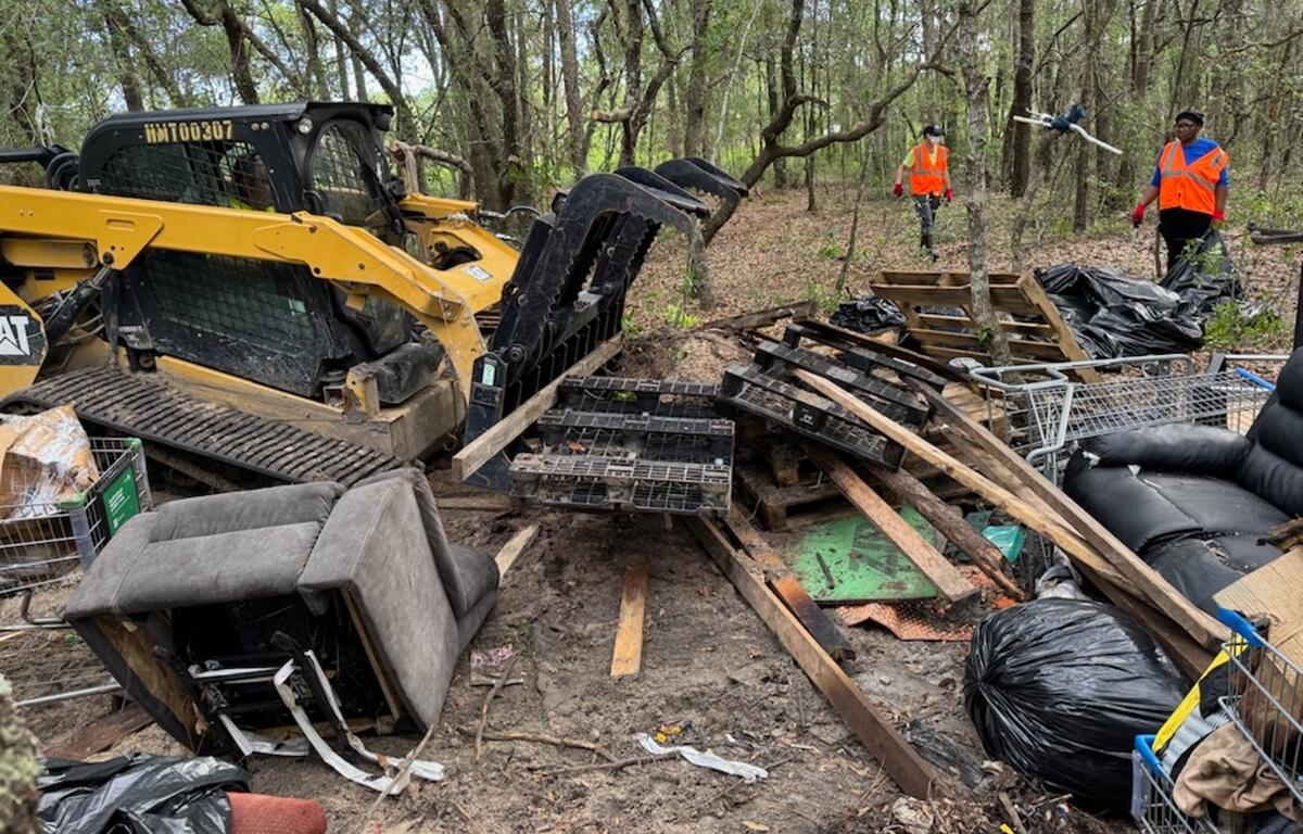 Bulldozer and workers clearing illegal dump site in wooded area, surrounded by trash, furniture, and shopping carts.