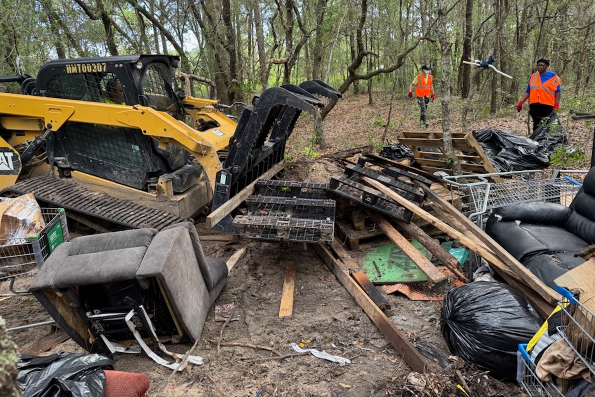 Bulldozer and workers clearing illegal dump site in wooded area, surrounded by trash, furniture, and shopping carts.