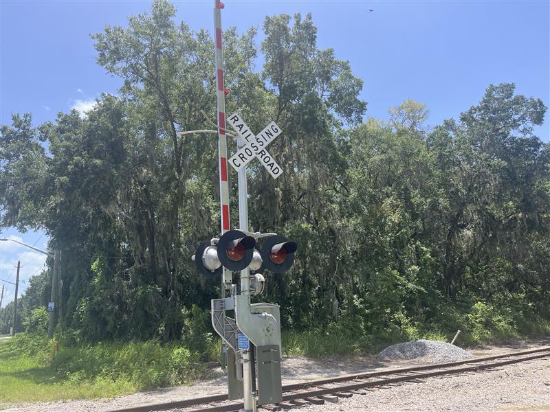 Railroad crossing gate and signal beside wooded tracks under a blue sky in Ocala.