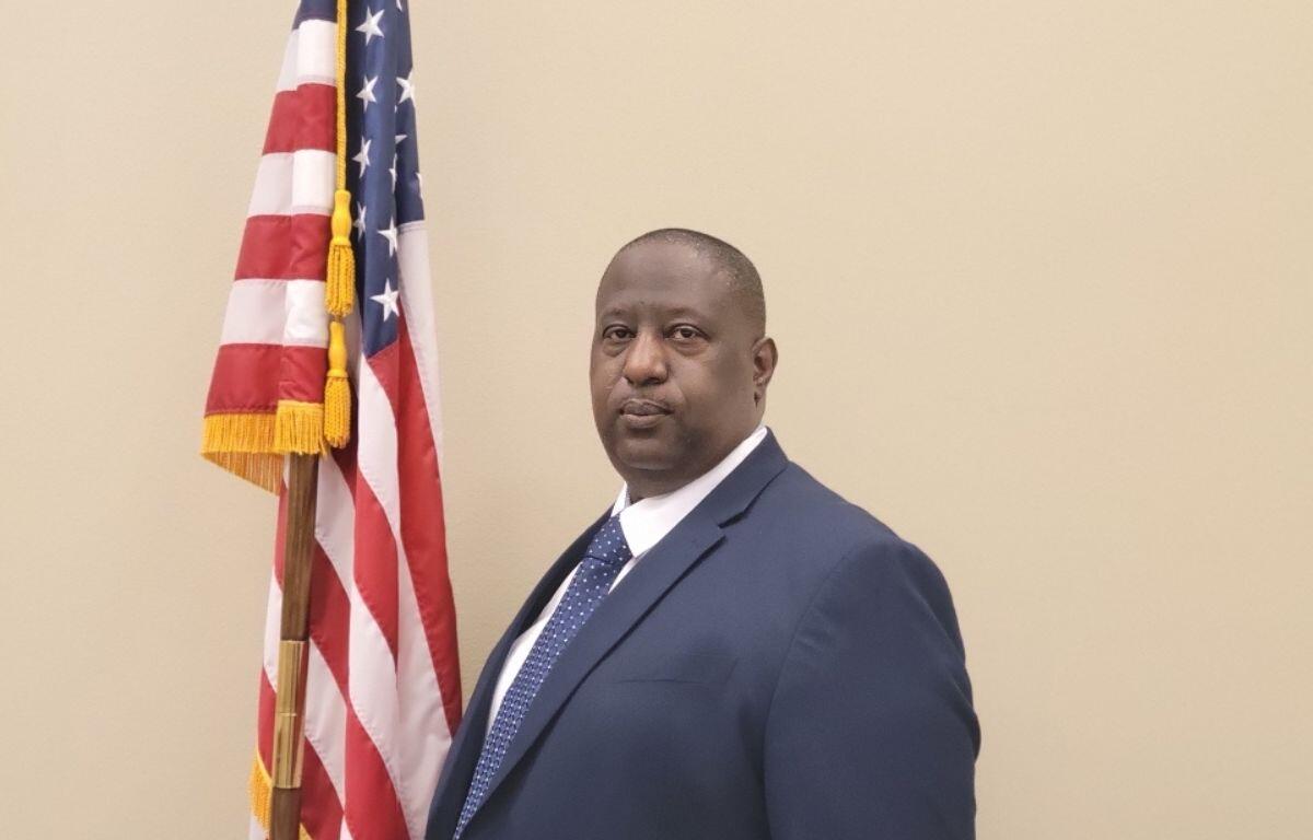 A photo of a man dressed in a suit, with the red, white and blue of the American flag to his left against a beige background.