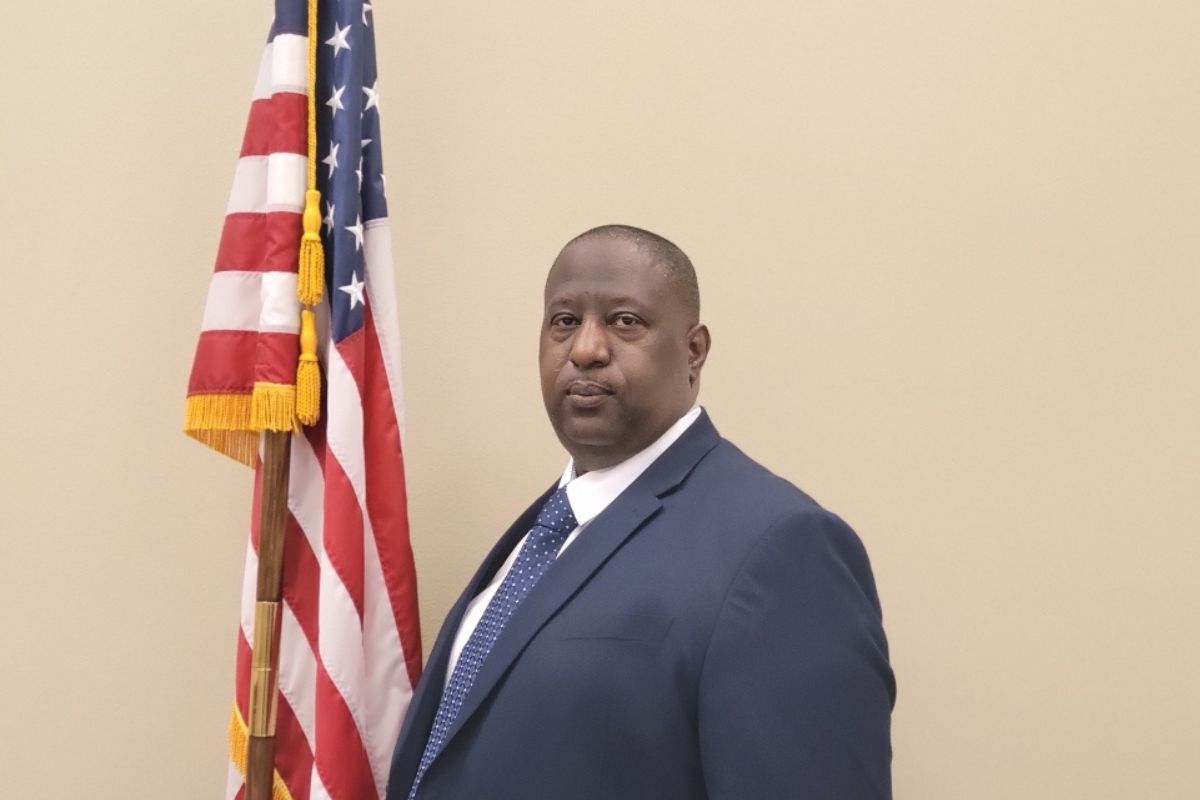 A photo of a man dressed in a suit, with the red, white and blue of the American flag to his left against a beige background.