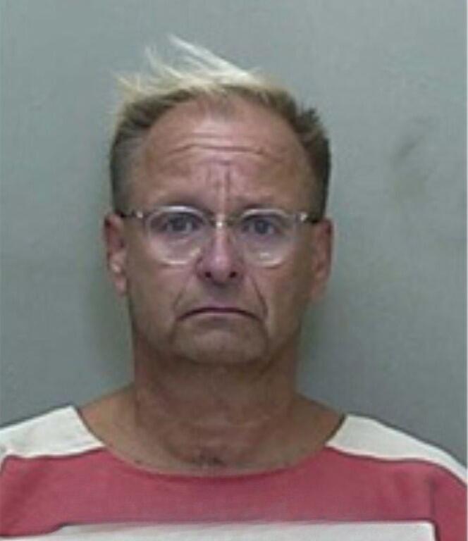 Mugshot of an older man with white hair and a mustache, wearing a red-and-white striped jail uniform.
