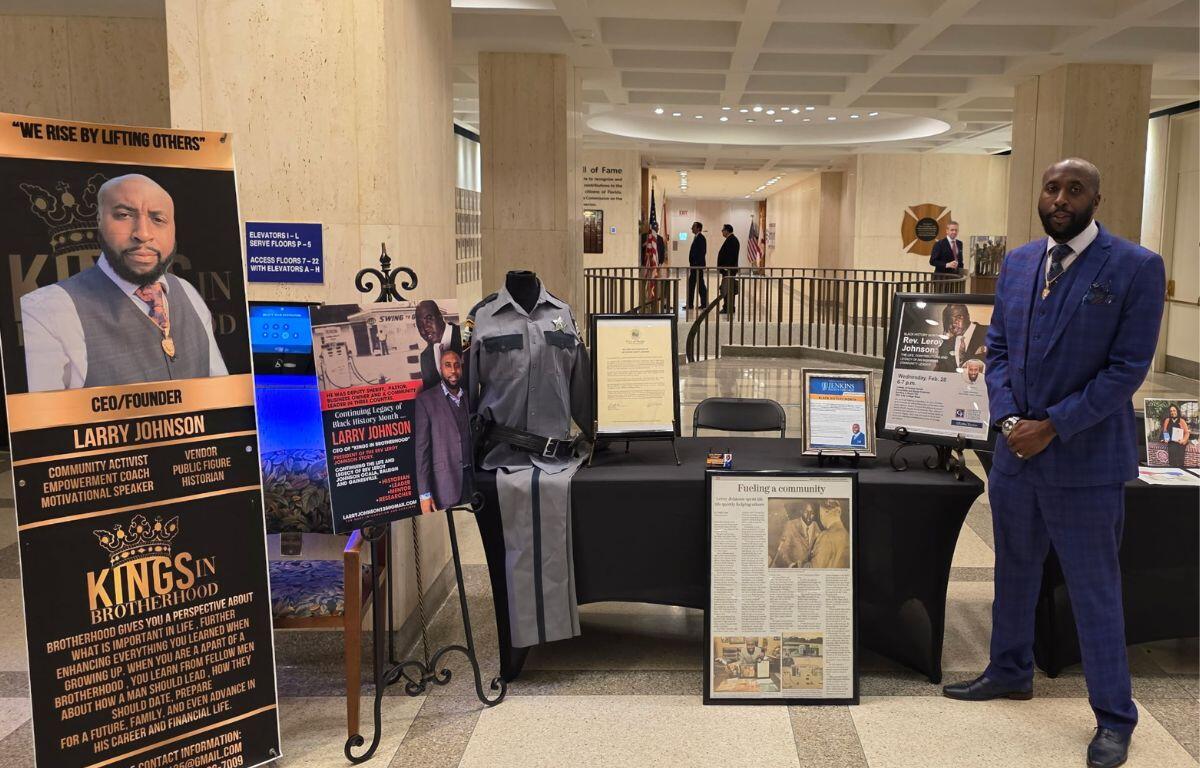 Several placards on stands, with writing a pictures to the left of a table, with various items on the table and in front of the table, with a man in a suit to to the right, with a deep background leading to a far corridor.