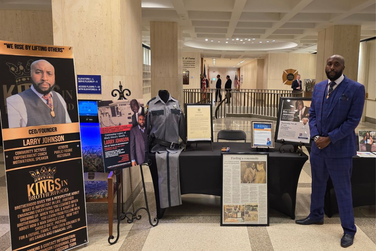 Several placards on stands, with writing a pictures to the left of a table, with various items on the table and in front of the table, with a man in a suit to to the right, with a deep background leading to a far corridor.