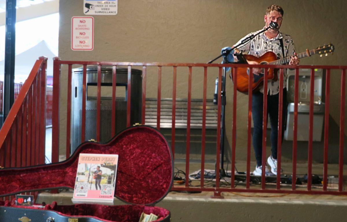 A solo performer playing guitar and singing on a small outdoor stage with speakers and a mic setup.