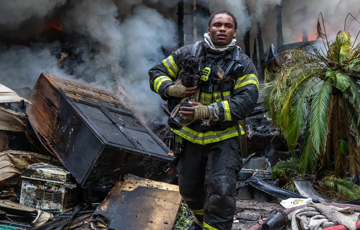 Firefighter carrying a dog away from a burning mobile home surrounded by smoke and debris.