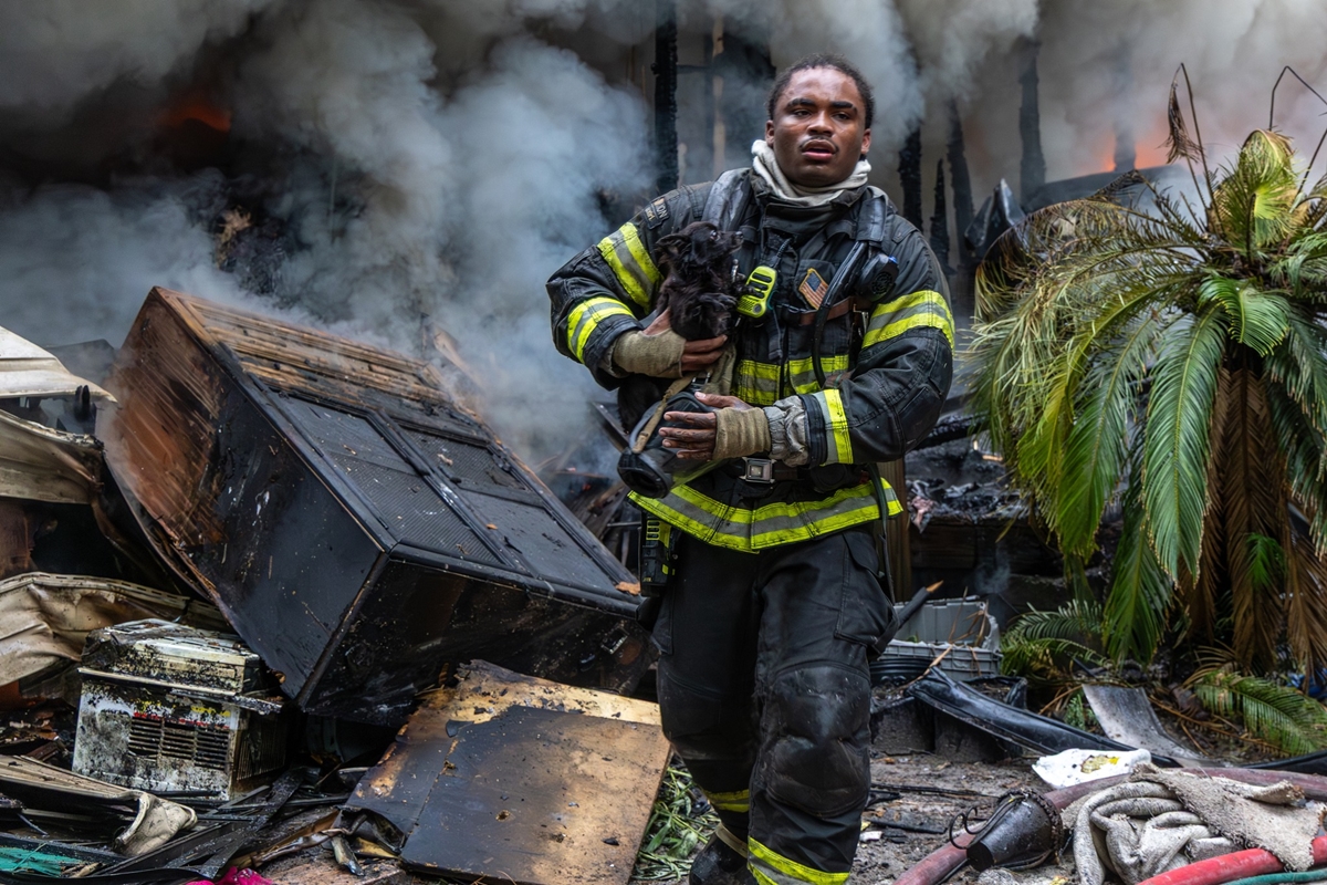 Firefighter carrying a dog away from a burning mobile home surrounded by smoke and debris.