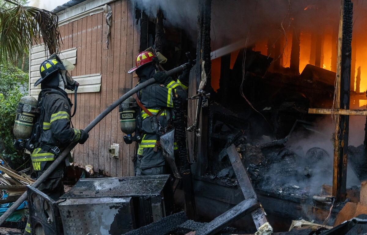 Two firefighters spray water into a burning mobile home as flames and smoke pour from inside.