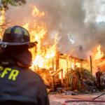 Firefighter watches a mobile home completely engulfed in flames with heavy smoke rising.