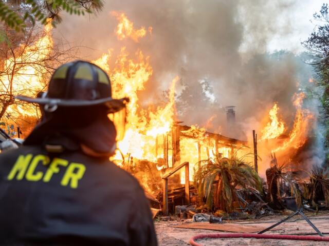 Firefighter watches a mobile home completely engulfed in flames with heavy smoke rising.