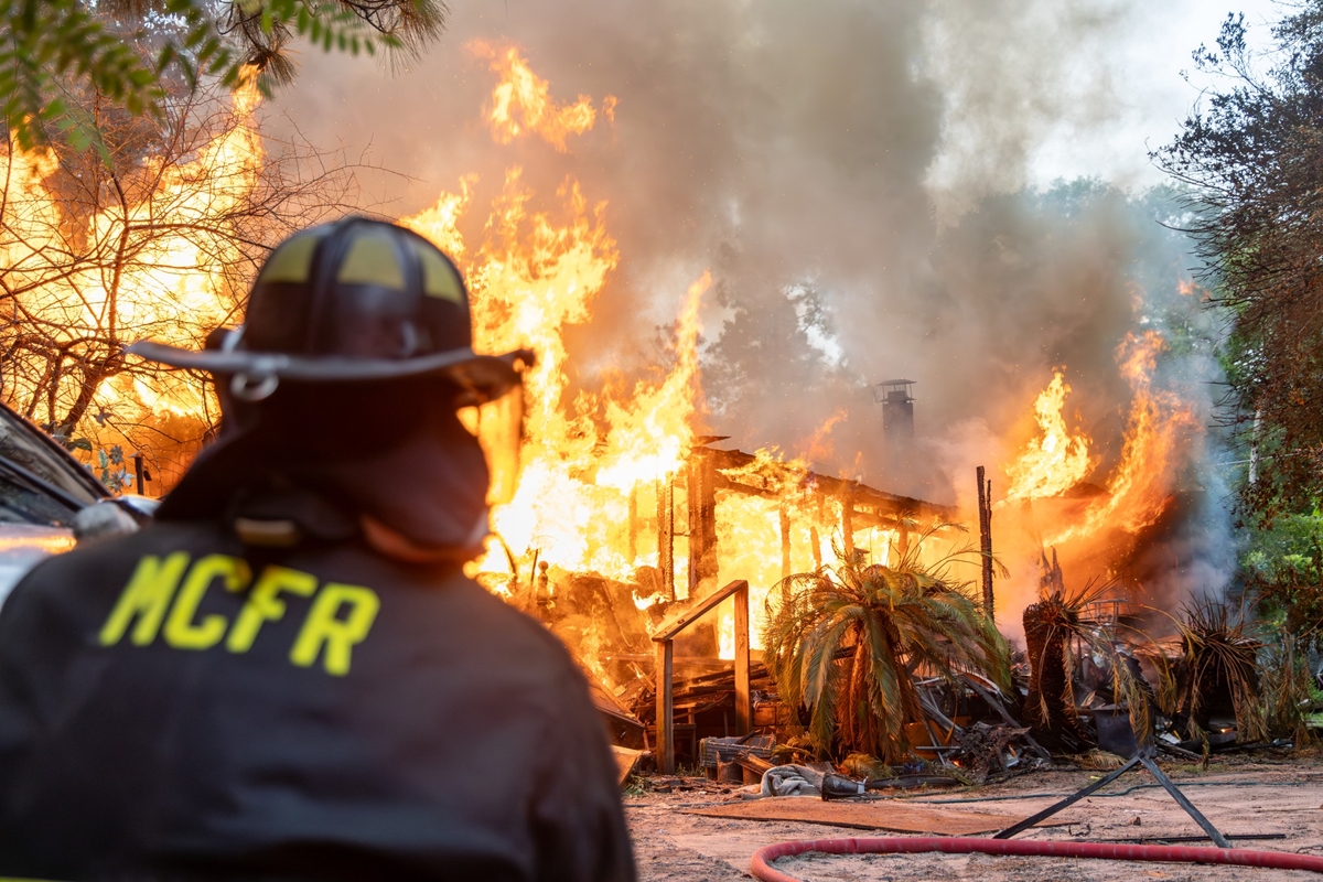 Firefighter watches a mobile home completely engulfed in flames with heavy smoke rising.
