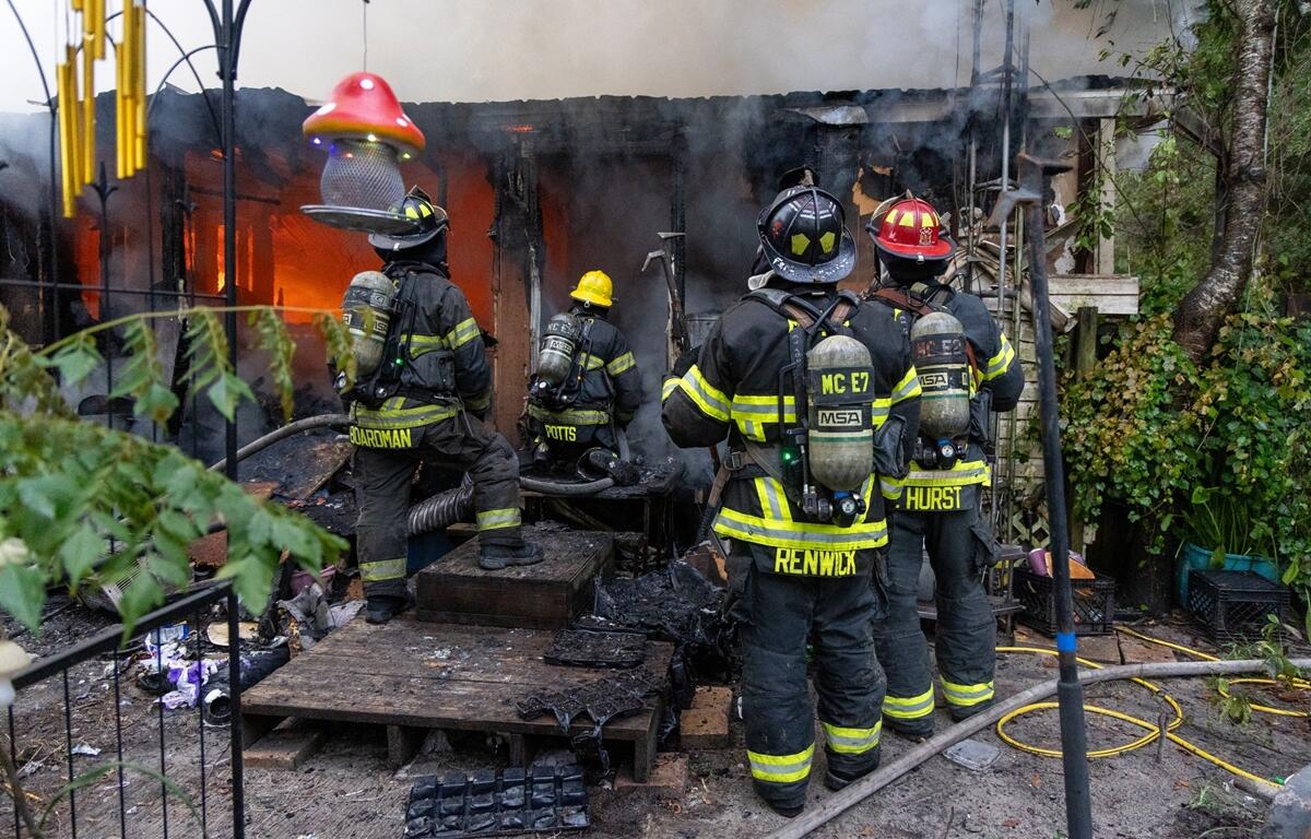 Group of firefighters in full gear stand in front of a burning mobile home with flames and smoke pouring from inside.