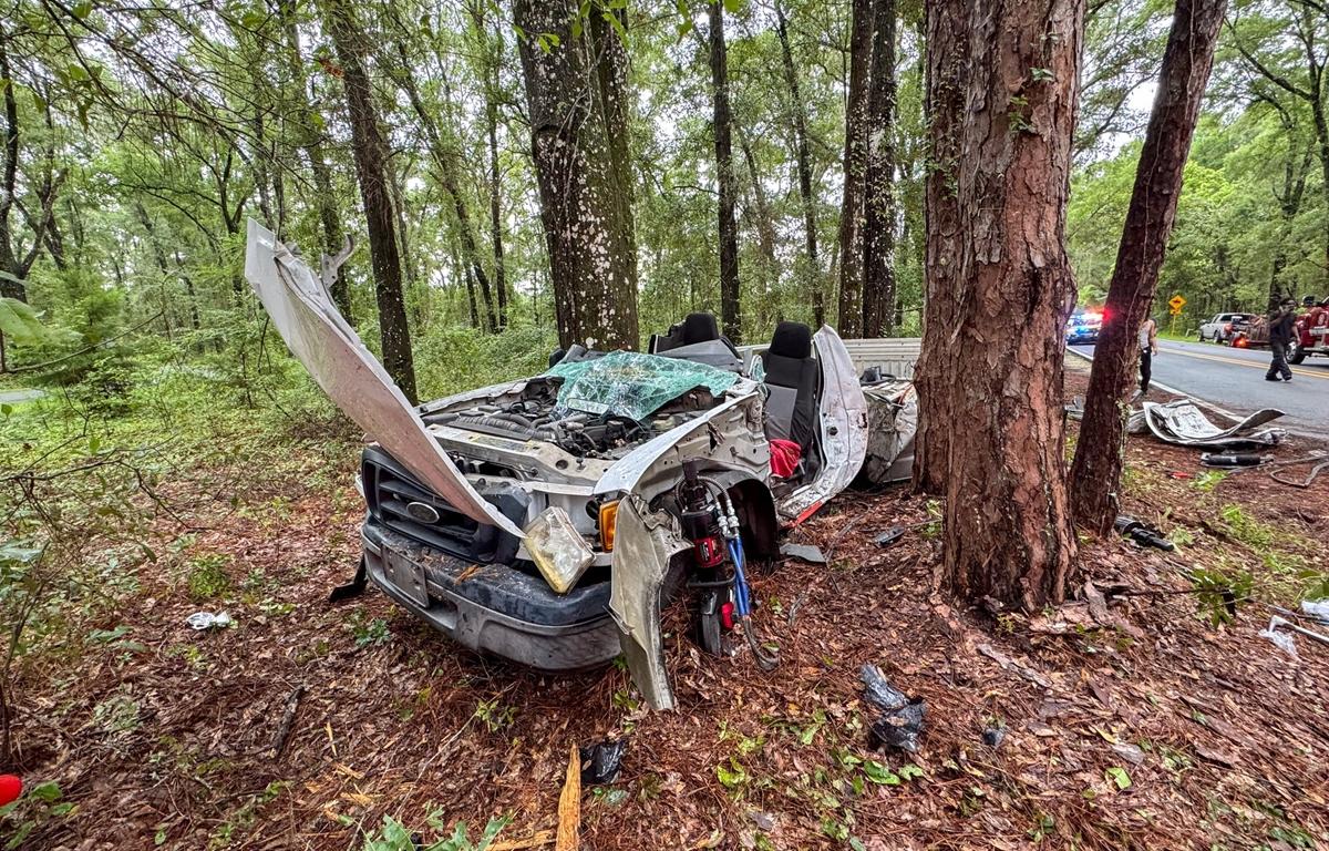 Severely damaged pickup truck with windshield shattered and hood open, resting against tree.