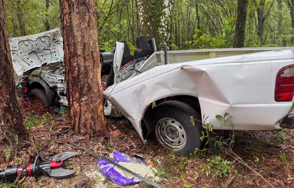 Crashed white pickup truck wrapped around trees in wooded area as fire rescue tools lie nearby.