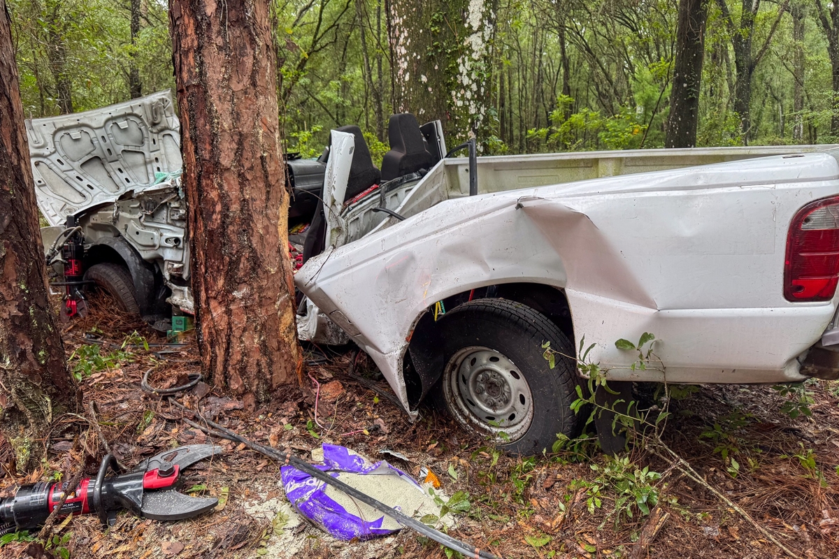 Crashed white pickup truck wrapped around trees in wooded area as fire rescue tools lie nearby.