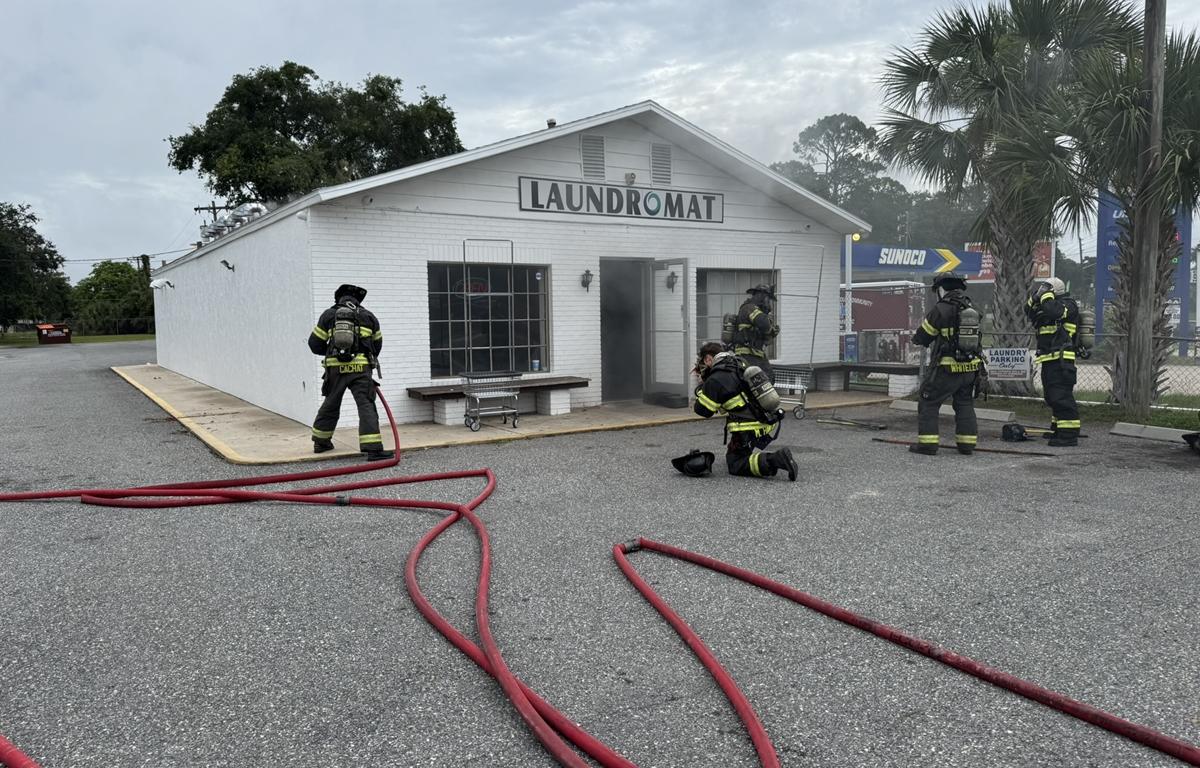 Firefighters outside a white laundromat with smoke coming from the doorway and hoses on the ground.