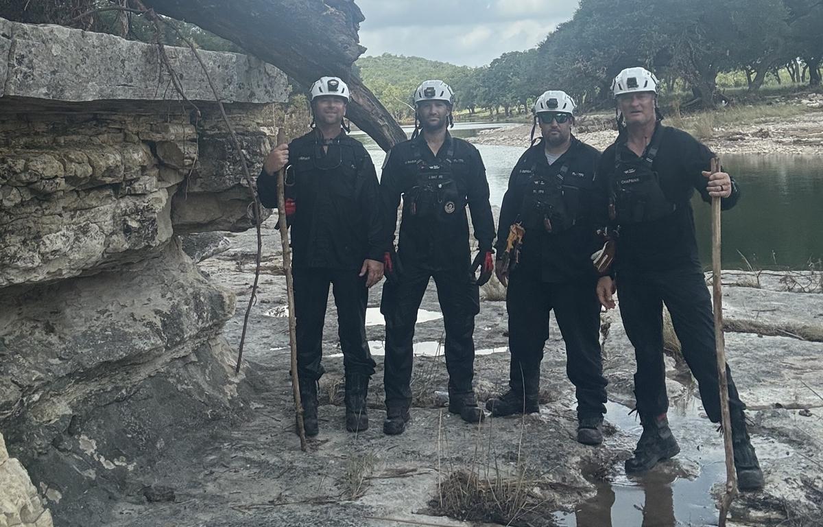 Four Task Force 8 members in black uniforms and helmets stand beside rocky riverbank in Texas