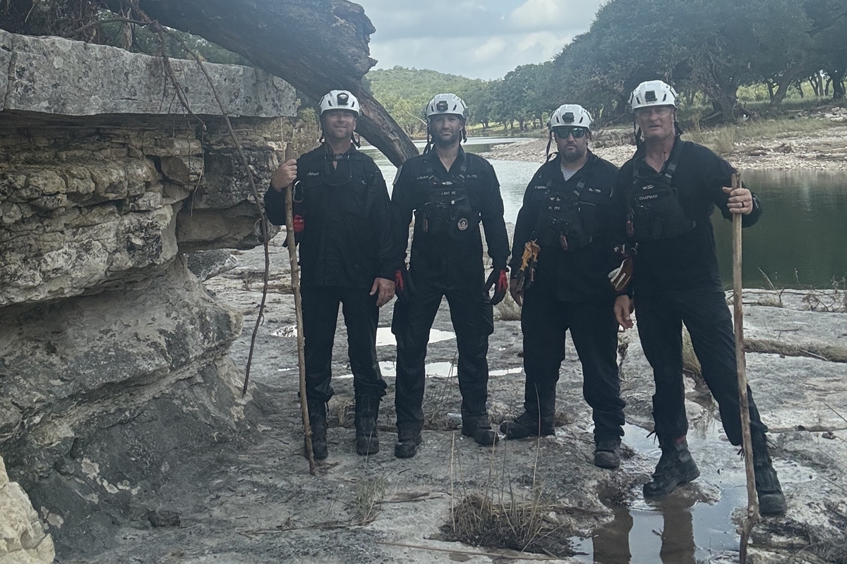 Four Task Force 8 members in black uniforms and helmets stand beside rocky riverbank in Texas