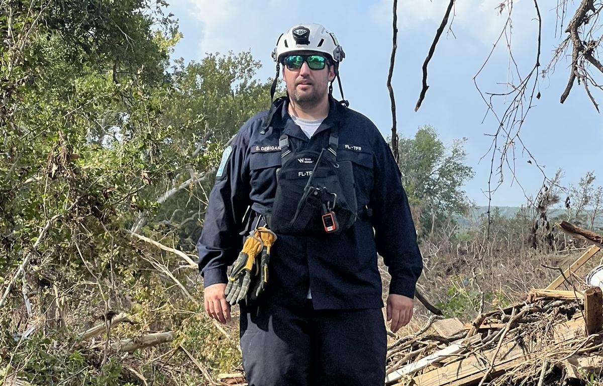 Florida Task Force 8 member wearing helmet and gear stands near flood debris in Texas