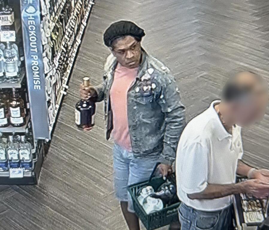 Surveillance image of a man holding a bottle of liquor and a green shopping basket inside a store.