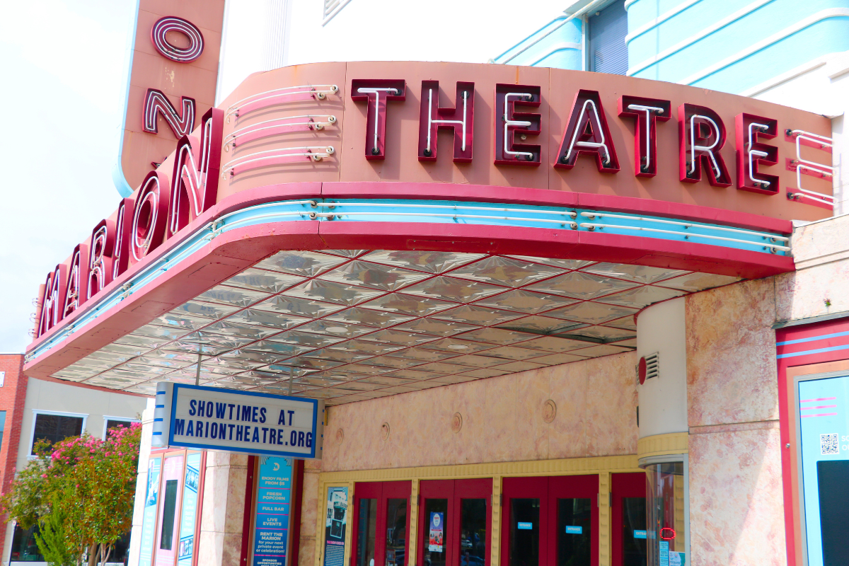 The exterior of the Marion Theatre, featuring a retro-style marquee with red neon letters and a sign below reading "Showtimes at MarionTheatre.org." The building has a pink and cream facade with red doors and art deco accents.