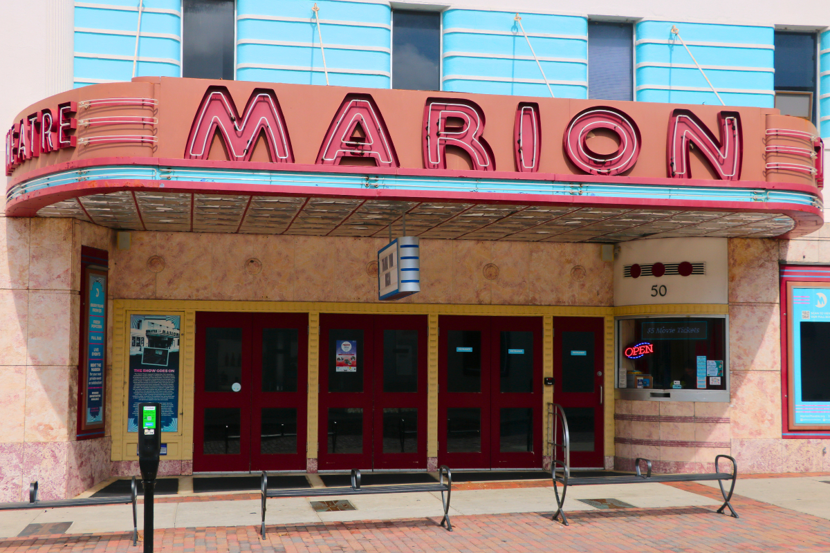 Front entrance of the Marion Theatre with a large, neon marquee sign above the red double doors displaying “MARION” in large red letters. Hanging from the marquee is a white and blue-striped sign. A ticket booth is situated on the right with a neon, “OPEN” sign. A parking meter and public benches are visible on the sidewalk in front.