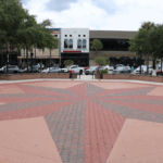 A wide, open town square with a red and gray brick star pattern in the center, surrounded by low brick walls and benches. In the background, cars are parked along the street in front of several storefronts. Trees line the sidewalk, providing shade, and flower planters add color to the scene.